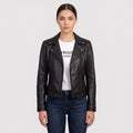 A woman modeling a black leather moto jacket over a white graphic t-shirt and dark blue jeans, shot against a solid light gray background.