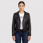 A woman modeling a black leather moto jacket over a white graphic t-shirt and dark blue jeans, shot against a solid light gray background.